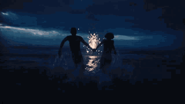 a man and a woman are holding hands in the ocean as fireworks are displayed in the background