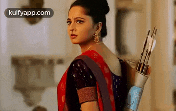 a woman in a red saree is holding a bow and arrow behind her back .