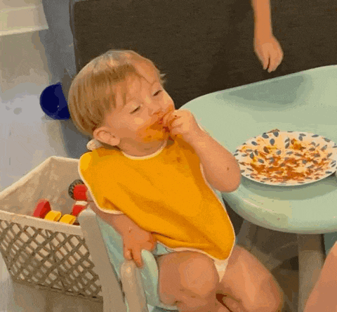 a baby wearing a yellow bib is sitting at a table eating food from a plate .