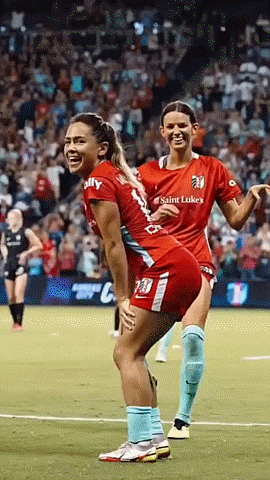two female soccer players wearing saint luke 's jerseys are having fun on the field