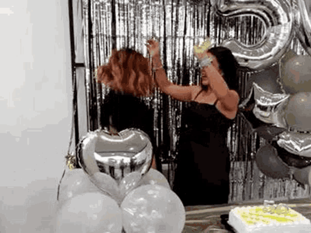 two women are dancing in front of a cake and balloons at a party .