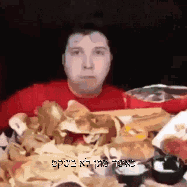 a man in a red shirt is sitting at a table with a bunch of food and the words in hebrew