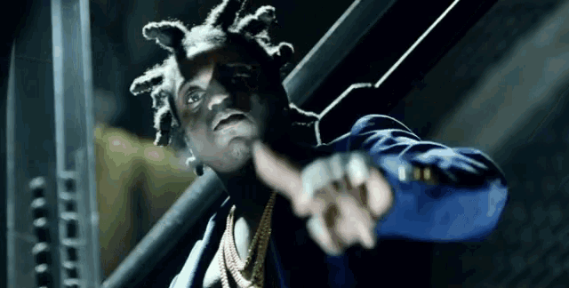 a man in a blue jacket and gold chains is pointing at the camera