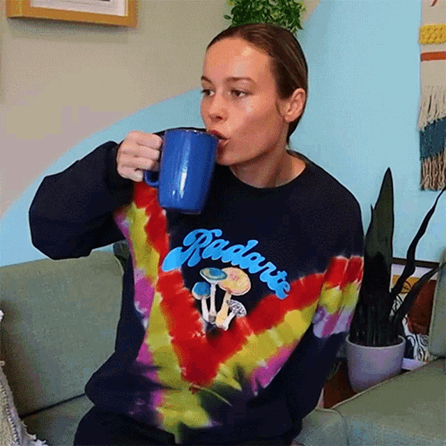 a woman wearing a tie dye sweatshirt is drinking from a blue mug ..