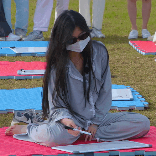 a woman wearing a mask and sunglasses sits on a mat holding a marker