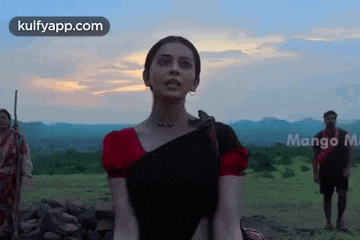 a woman in a black saree is standing in a field with a man in a red shirt behind her .