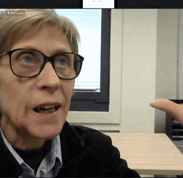 a woman wearing glasses and a black jacket is pointing at something in front of a screen that says removed