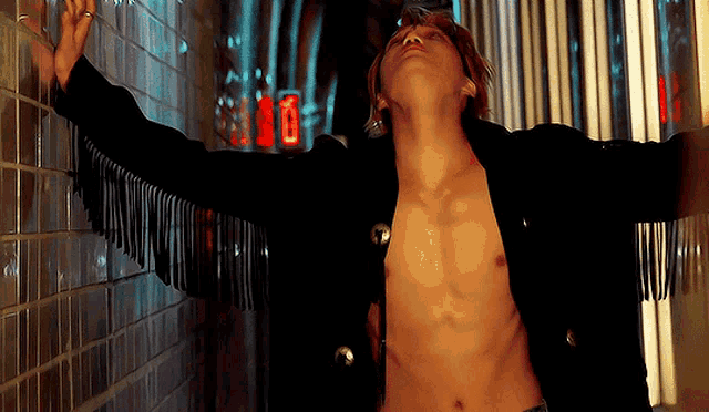 a shirtless man leans against a wall with his arms outstretched and looks up at the sky
