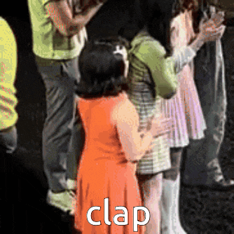 a little girl in an orange dress is standing in front of a group of people and claps .