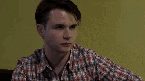 a young man in a plaid shirt is sitting at a table looking down .
