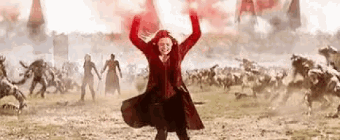 scarlet witch is standing in a field with her arms in the air and a crowd of people behind her .