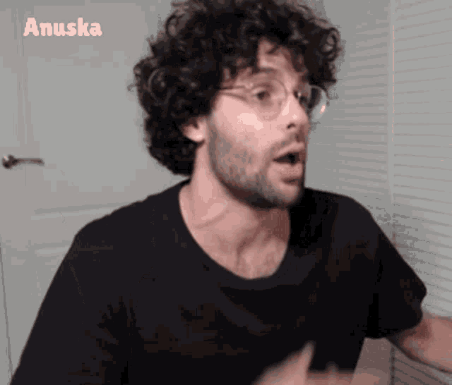 a man with curly hair and glasses is wearing a black shirt and the name anuska is on the bottom