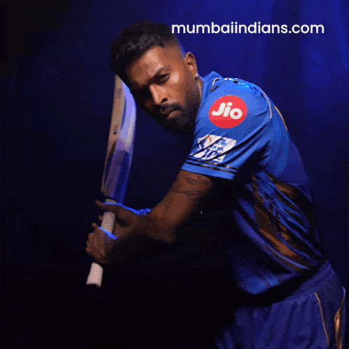 a man in a blue shirt is holding a bat and the website mumbaiindians.com is visible behind him