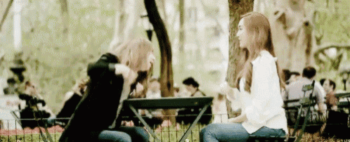 two women are sitting at a table in a park talking .