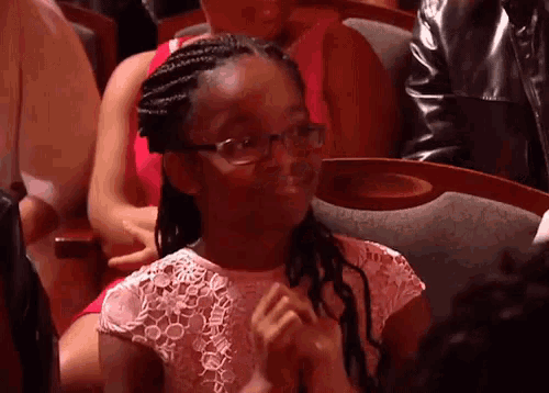 a young girl wearing glasses sits in a theatre with her hands folded