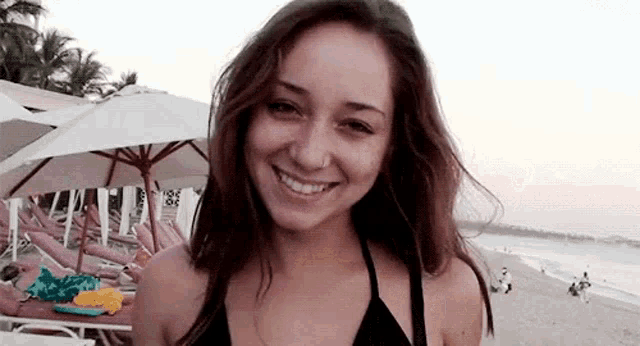 a woman in a black bikini is smiling at the camera on the beach .