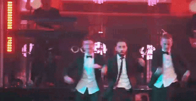 a group of men in tuxedos and bow ties are dancing on a stage in a dark room .