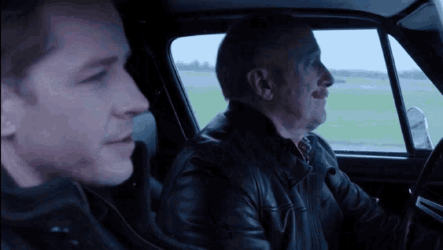 two men in leather jackets are sitting in a car looking out the window