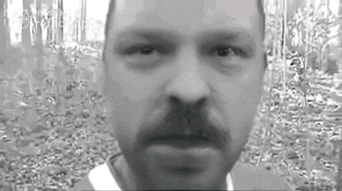 a black and white photo of a man with a mustache standing in the woods .