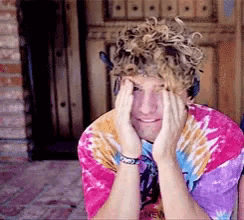 a man with curly hair is wearing a tie dye shirt and covering his face with his hands