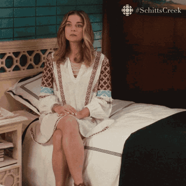 a woman sits on a bed with a #schitts creek logo in the corner