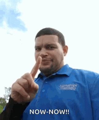 a man in a blue shirt is pointing his finger at the camera and saying `` now-now ! ''