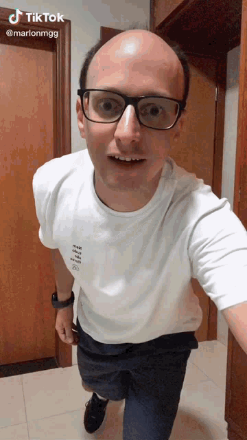 a bald man wearing glasses and a white t-shirt has a tiktok sticker above his head