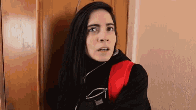 a woman wearing a black jacket and a red backpack is standing in front of a wooden door