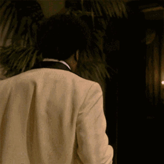 a man in a tuxedo is walking in a dark room with a palm tree in the background