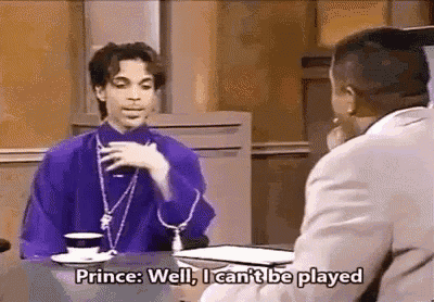prince is sitting at a table talking to a man while holding a cup of coffee .