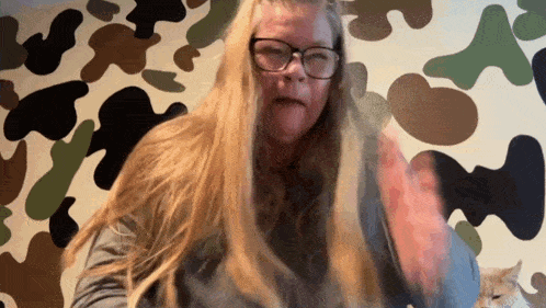 a woman with long blonde hair wearing glasses is standing in front of a camouflage wall