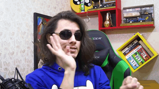 a man wearing sunglasses and a racer sweatshirt holds up his hand