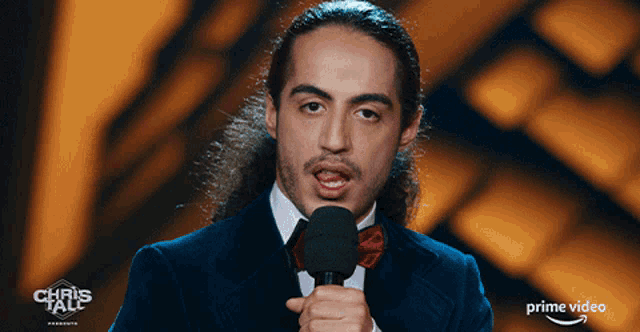 a man in a suit and bow tie is speaking into a microphone in front of a sign that says chris tall