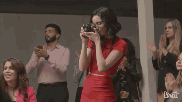 a woman in a red dress is taking a picture with a camera and the word brat is on the bottom right