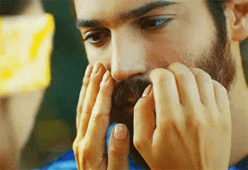 a man with a beard is holding his hands to his face and looking down .