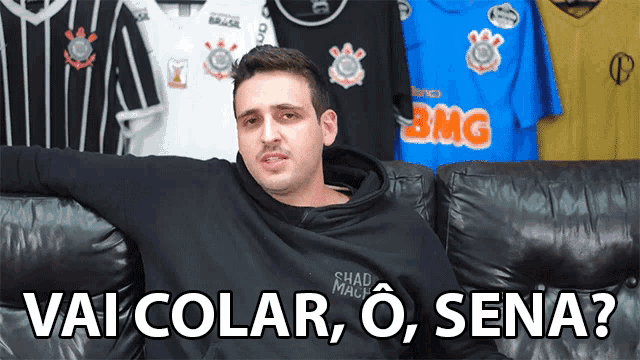 a man is sitting on a couch in front of a wall of shirts with one that says bmg on it