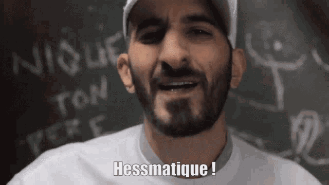 a man with a beard says hessmatique in front of a chalkboard