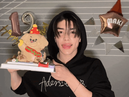 a man wearing a black aders sweatshirt holds a cake with a teddy bear on it