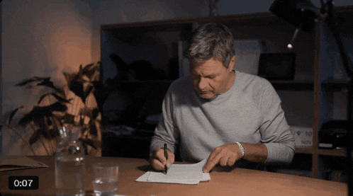a man sits at a table writing on a piece of paper with 0:07 on the bottom of the screen
