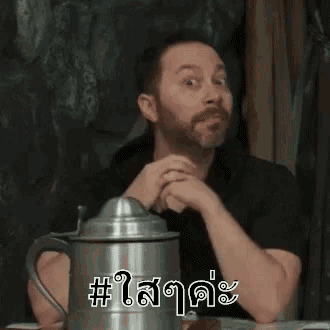 a man with a beard is sitting at a table next to a metal pitcher .