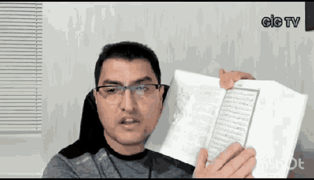 a man wearing glasses is holding a book in front of a window .