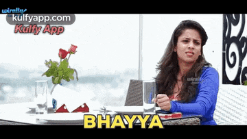 a woman sits at a table with a vase of flowers and says ' bhavya ' on the screen