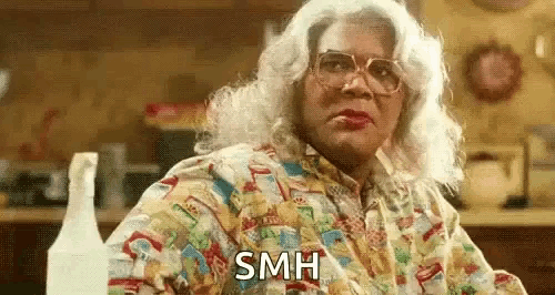 a woman in a wig and glasses is sitting at a table in a kitchen and says `` smh '' .