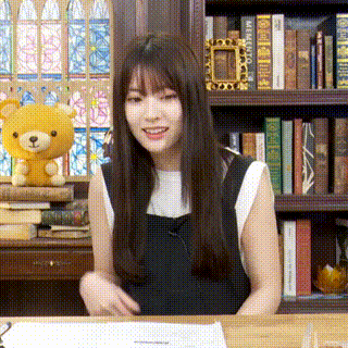a woman is sitting at a desk in front of a bookshelf with a teddy bear in the background .