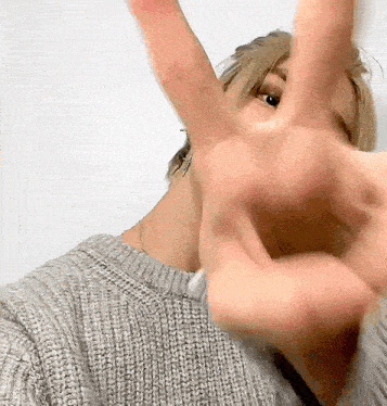 a woman wearing a grey sweater is making a peace sign with her hand .