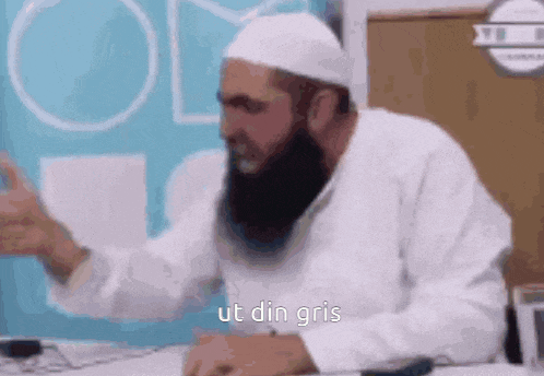 a man with a beard and a white hat is sitting at a desk with his arms outstretched .