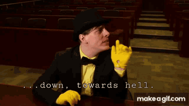 a man in a black suit and yellow gloves is sitting at a table and says " down towards hell "