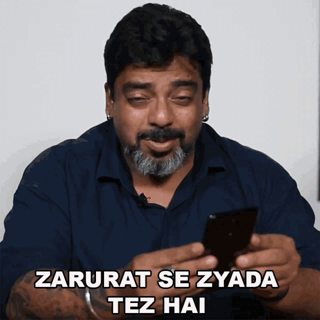 a man with a beard is holding a cell phone and says " zarurat se zyada tez hai "