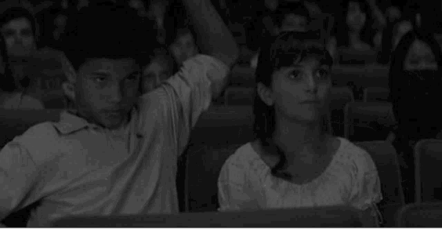 a boy and a girl are sitting in a theater watching a movie . the boy is raising his hand to answer the question .