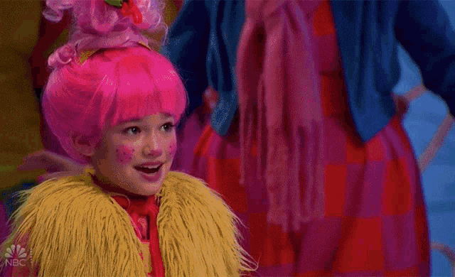 a girl in a pink wig and a yellow fur coat is standing in front of a sign that says nbc
